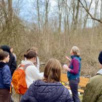 Careerline Tech Students standing with their backs to the camera, a teacher gesturing to trees behind herself, outside in cold weather next to a forest